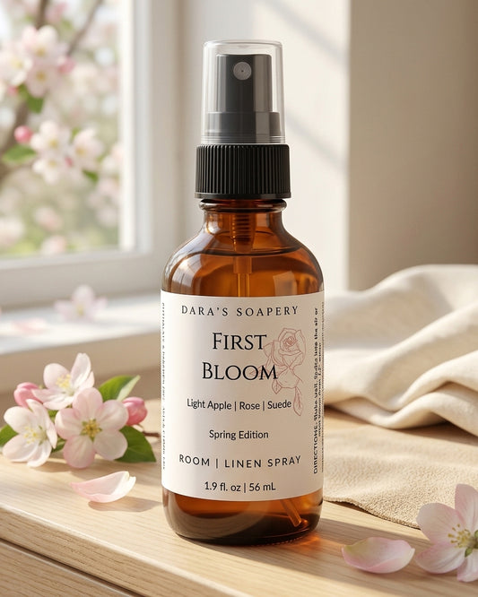 A 1.9 oz amber glass bottle of First Bloom Room and Linen Spray by Dara's Soapery sitting on a sunlit wooden table. The label features a rose illustration and scent notes of Light Apple, Rose, and Suede. The background includes fresh pink apple blossoms and soft cream linen in a bright, airy setting.