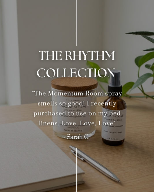 A candle and a bottle of 'The Rhythm Collection' room spray on a wooden surface with a plant and notebook in the background.